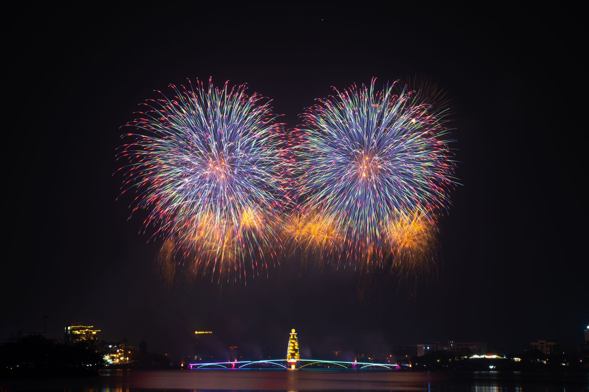 Brilliant fireworks illuminate the night sky over a vibrant cityscape with a lit bridge and waterfront.