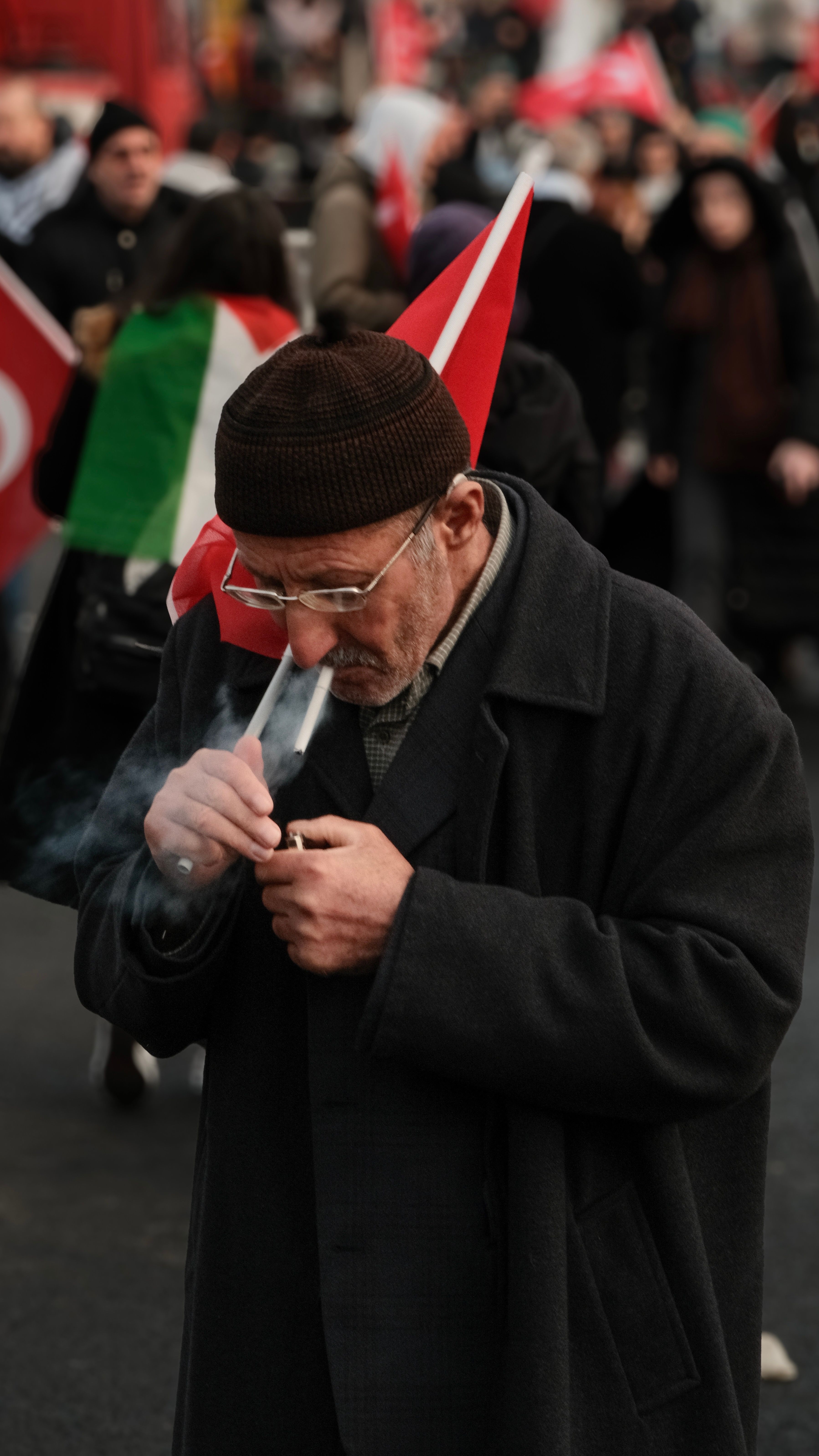 Senior adult man in coat lighting cigarette during a protest rally, flags in the background.
