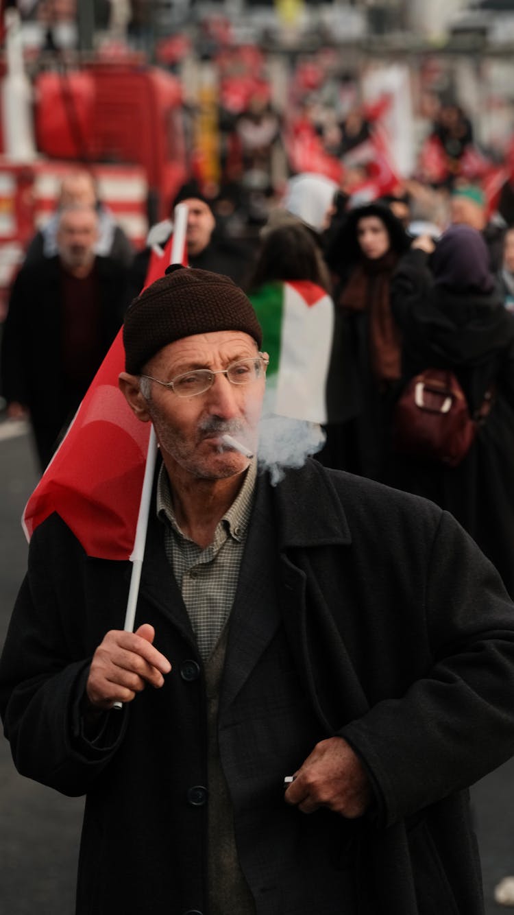 Elderly Man Smoking A Cigarette Walking In The Crowd And Holding A Flag 