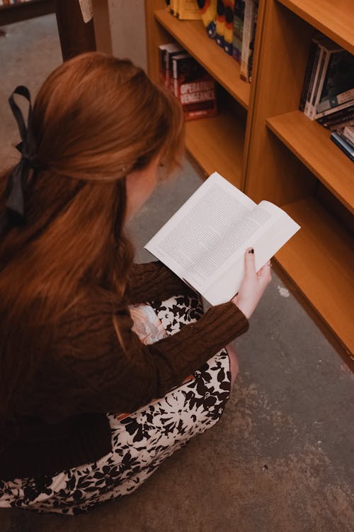 Redhead Woman Squatting and Reading Book in Library