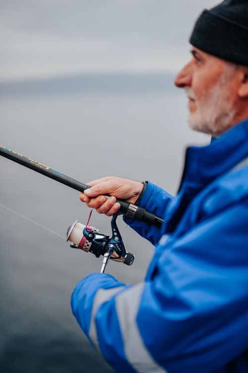 Free Elderly man enjoys fishing with a rod by the water, wearing a blue jacket and beanie. Stock Photo