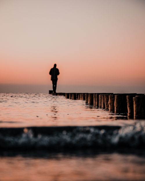 Free A silhouette of a person standing on a pier by the seashore at sunset, creating a tranquil scene. Stock Photo