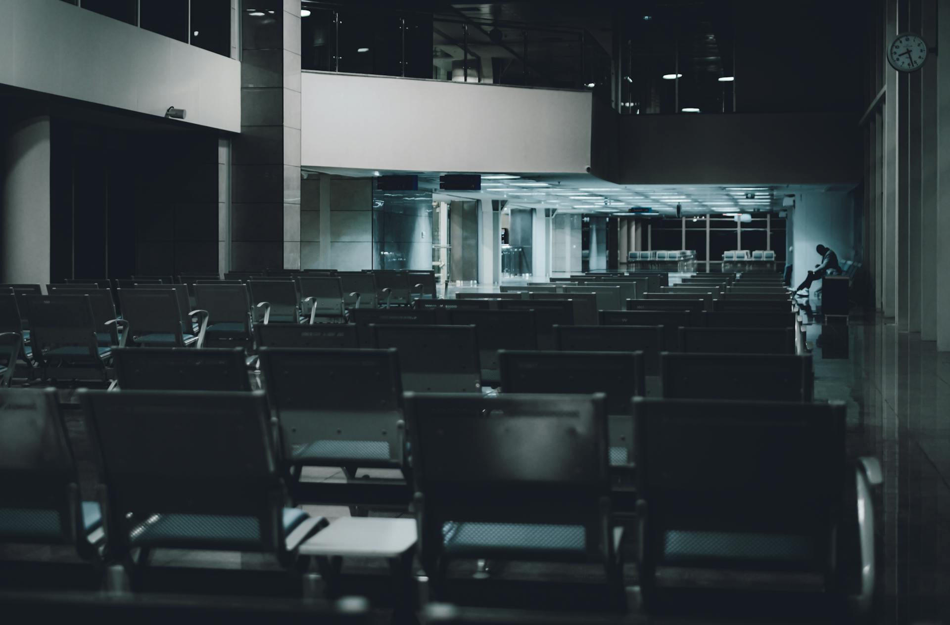 A quiet, dimly lit airport waiting area with unoccupied rows and a lone figure in the distance.