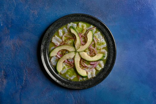 Top view of aguachile with avocado, onions on a black plate against blue background.