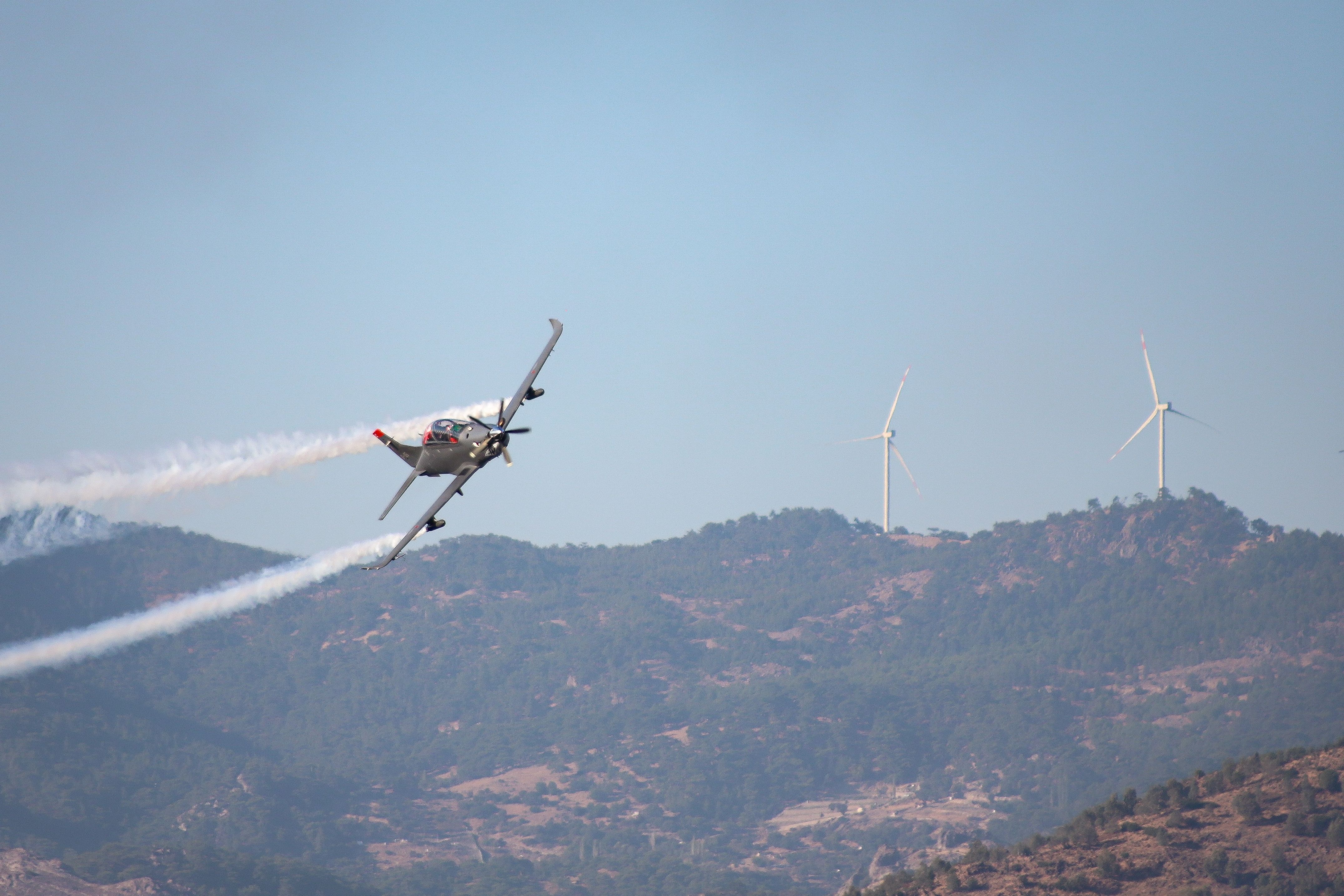 Aerobatic plane with smoke trail flying over the mountains with wind turbines in İzmir, Türkiye.