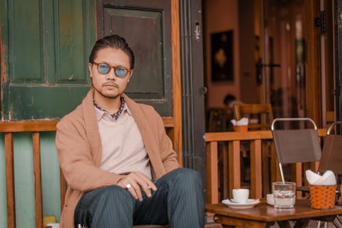 Man in Brown Cardigan and Blue Eyeglasses Sitting at a Cafe Table