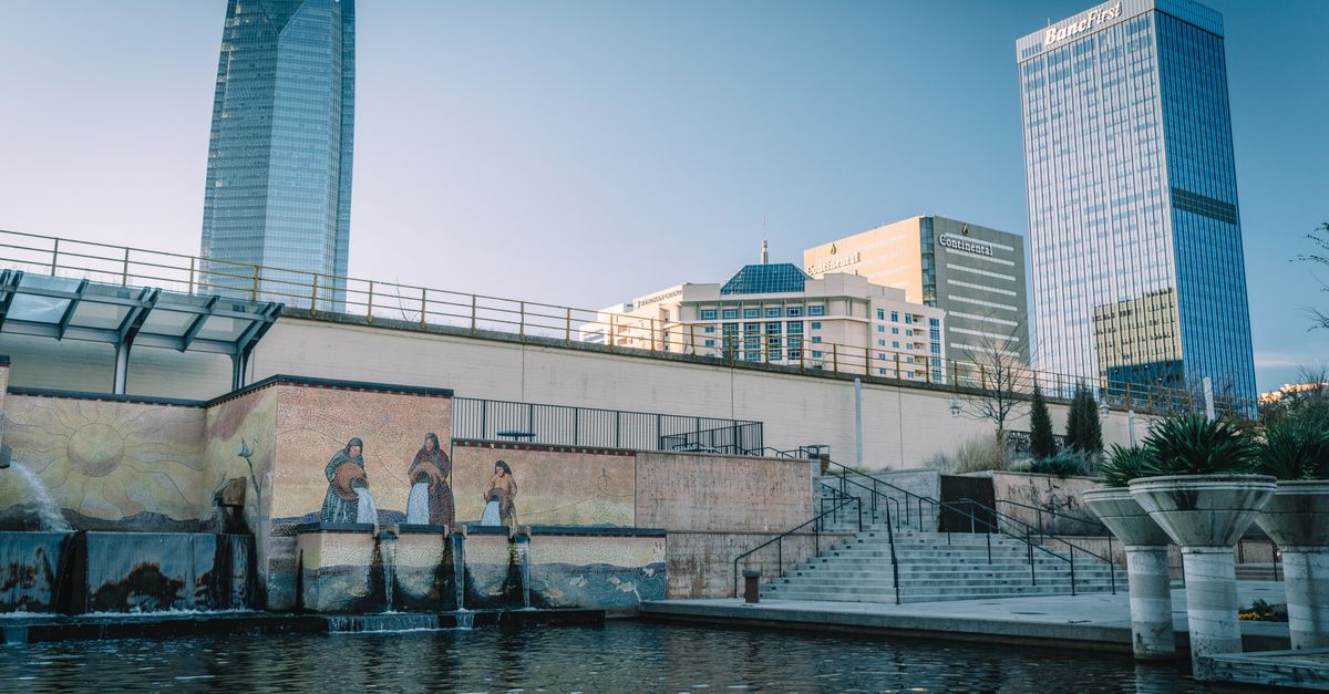 Cityscape featuring skyscrapers, a mural, and water feature with clear blue skies.