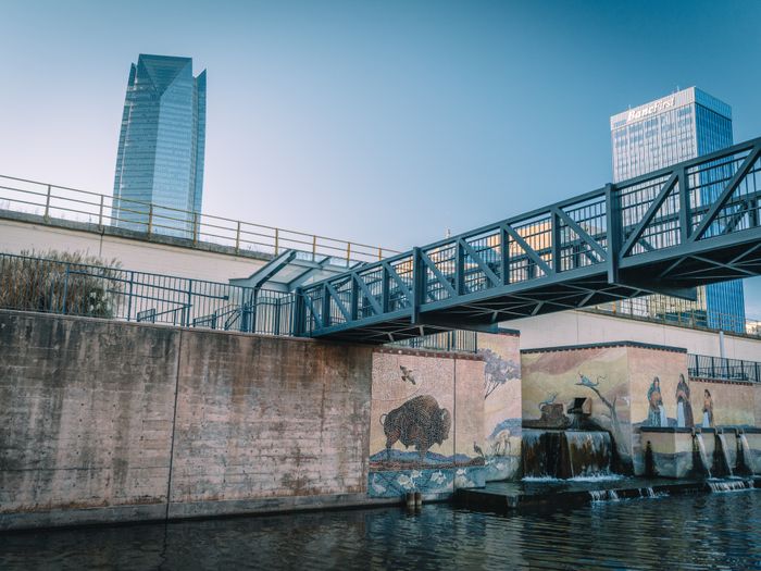 View of Oklahoma City's canal featuring a footbridge, murals, and skyscrapers in the background.
