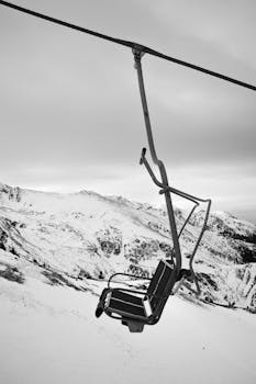 Black and white photograph of an empty ski lift over snow-covered mountains, capturing a serene winter landscape.