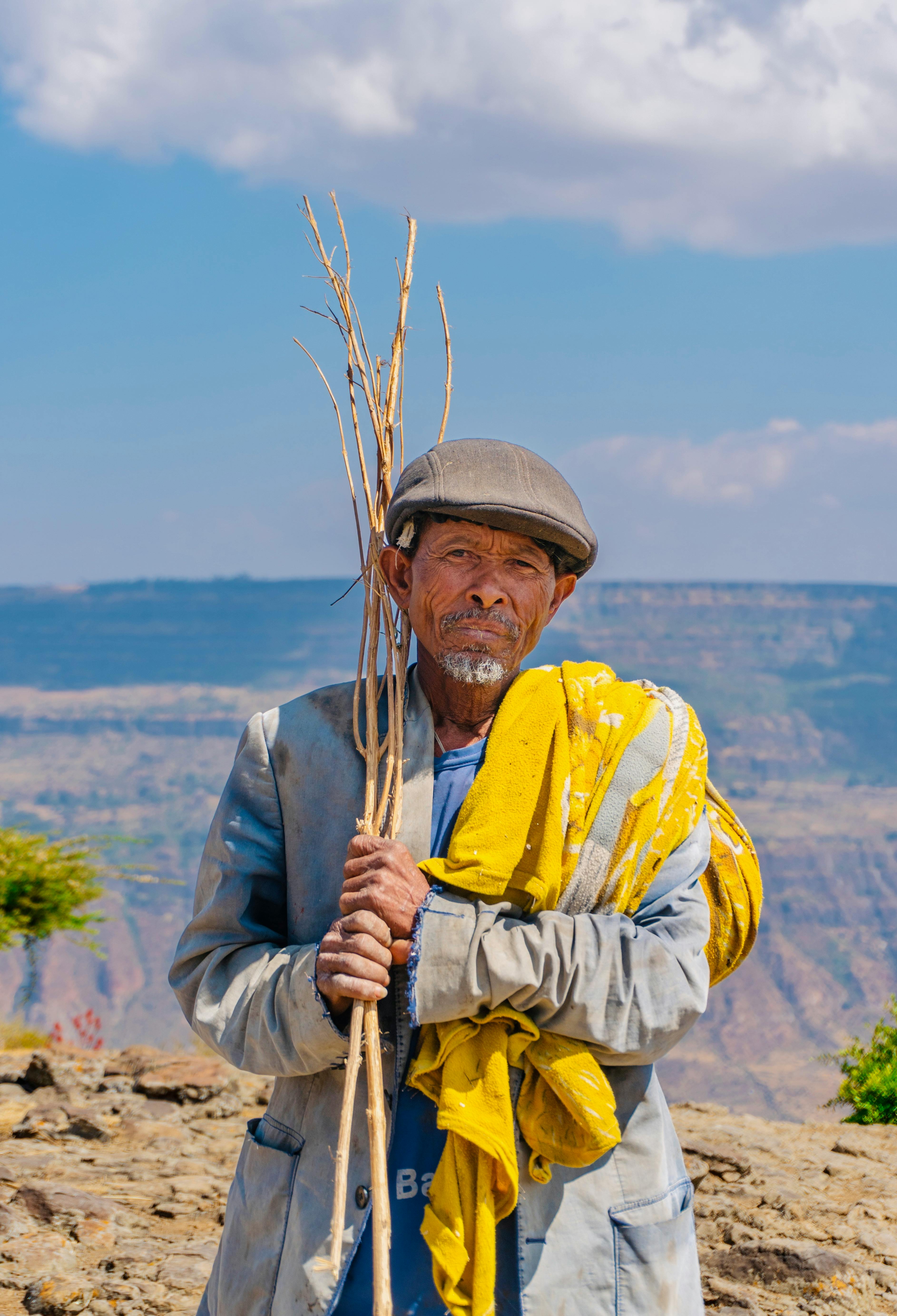 An elderly Ethiopian man holding sticks, standing in a scenic rural area under a clear blue sky.