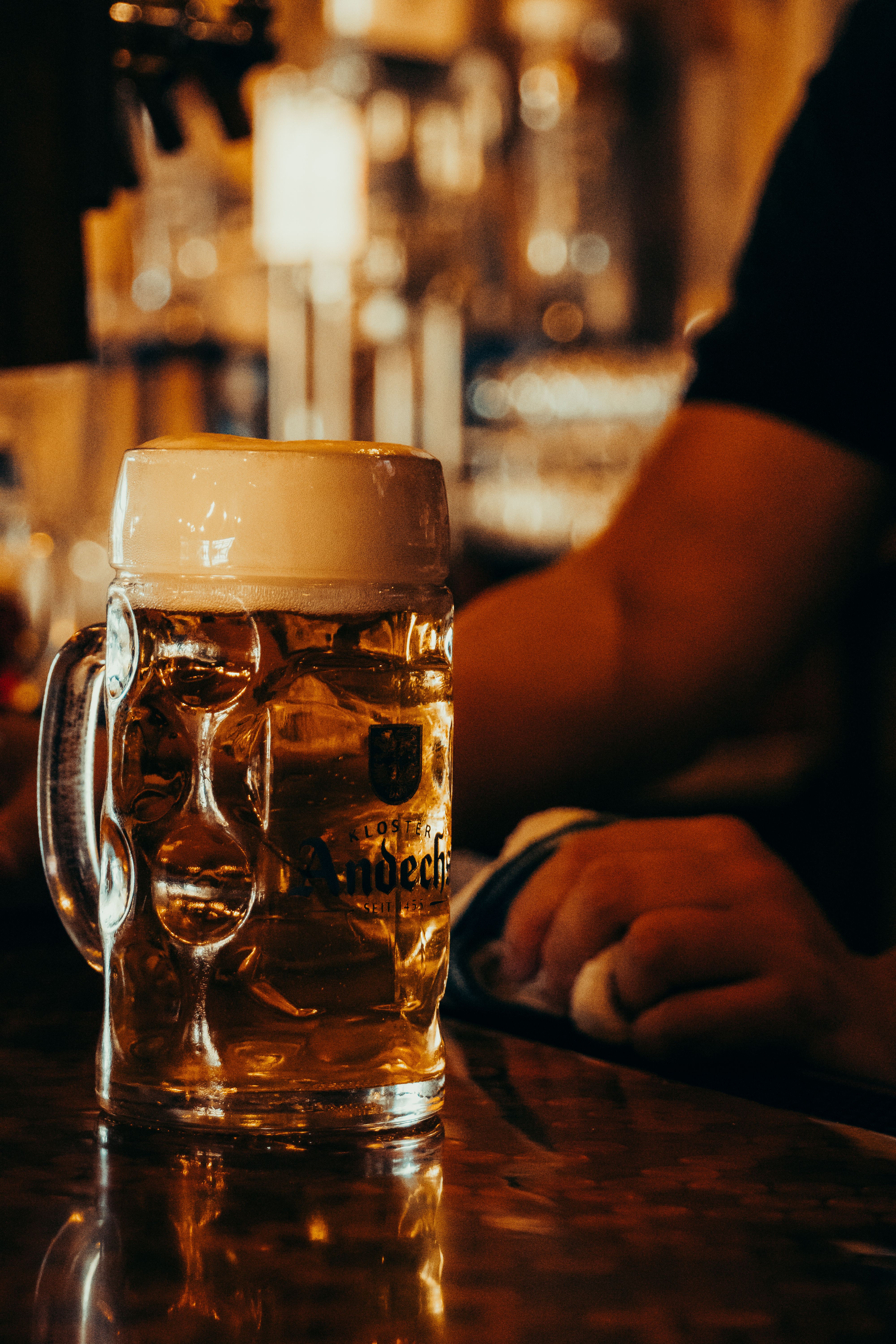 A frothy pint of beer on a bar counter with warm ambient lighting and a hand holding a towel.