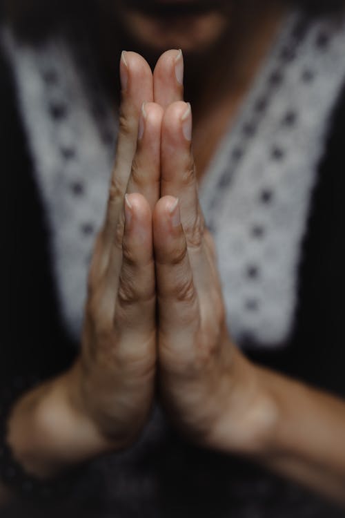 Close-up of woman praying or meditating with her hands together in namaste gesture and body blurred in the background on a dark moody atmosphere. Concept of faith, religion and belief in god