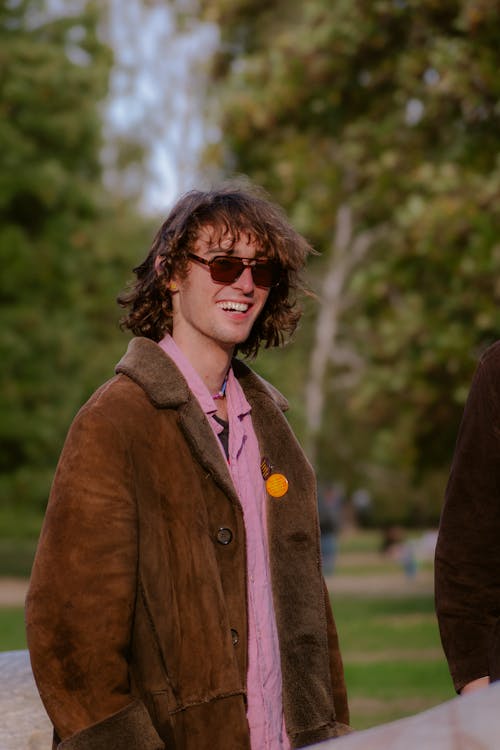 Man in a Brown Jacket and Sunglasses Standing in a Park and Smiling 