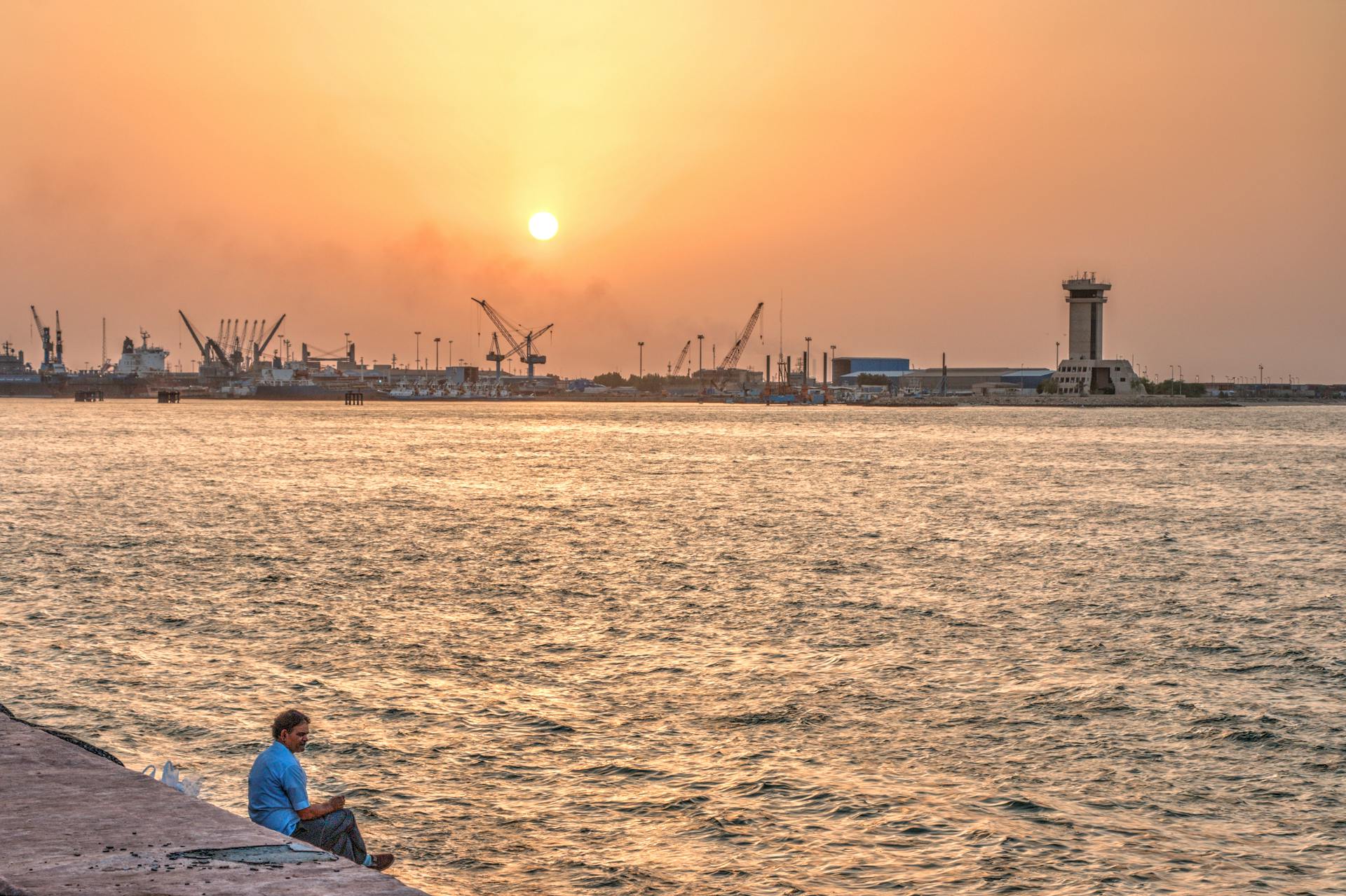 A peaceful sunset over an industrial harbor with cranes and sea, featuring a lone man on the pier.