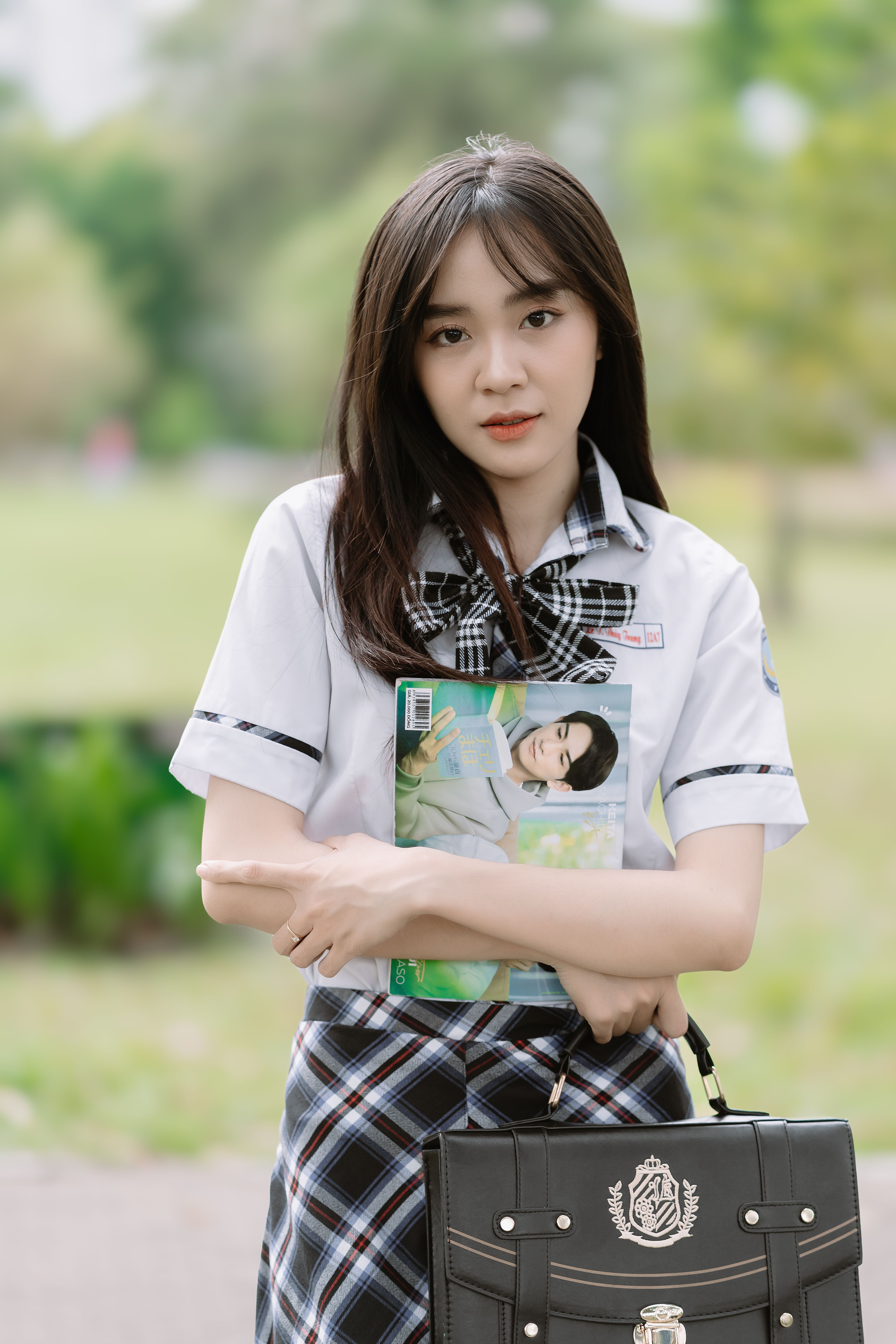 Elegant Asian woman in a school uniform standing outdoors, holding a magazine, embodying youth and sophistication.