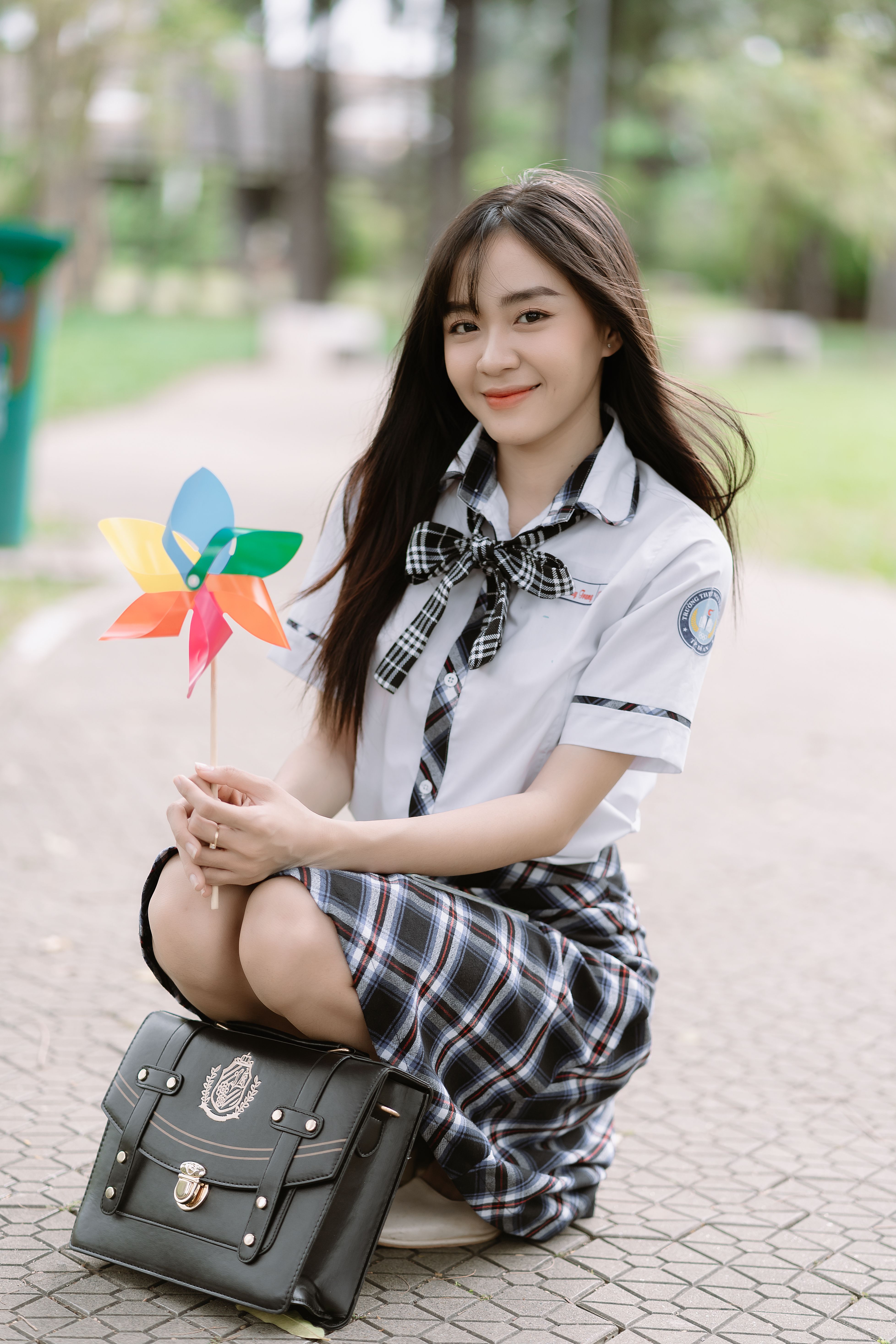 Smiling young woman holding pinwheel, wearing school uniform, squatting on pavement outdoors.