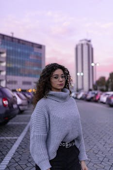 Stylish lady with curly dark hair in sweater looking at camera on parking at twilight