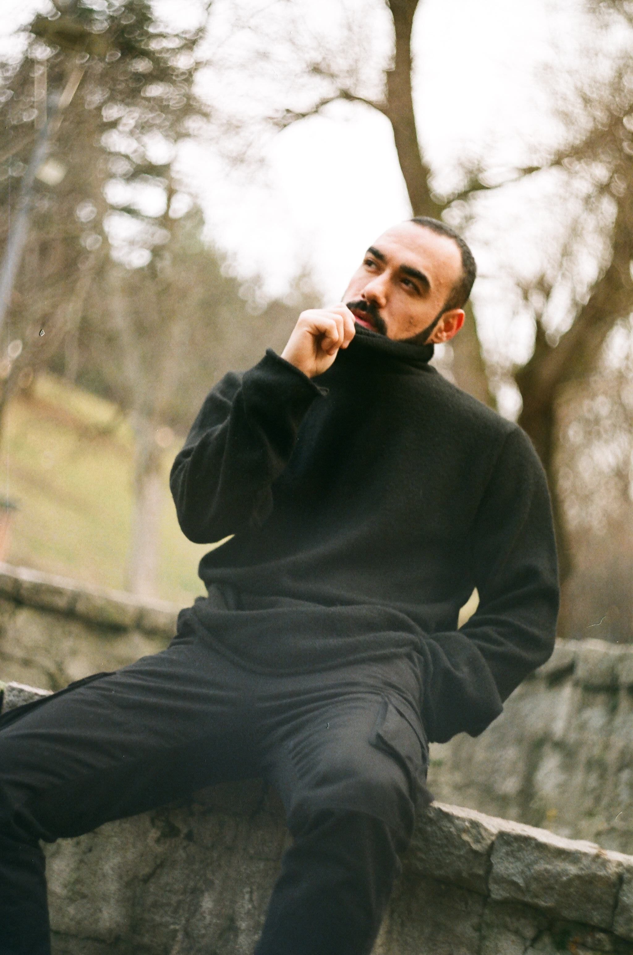 A man in a black pullover sits thoughtfully outdoors. Fashionable and pensive.