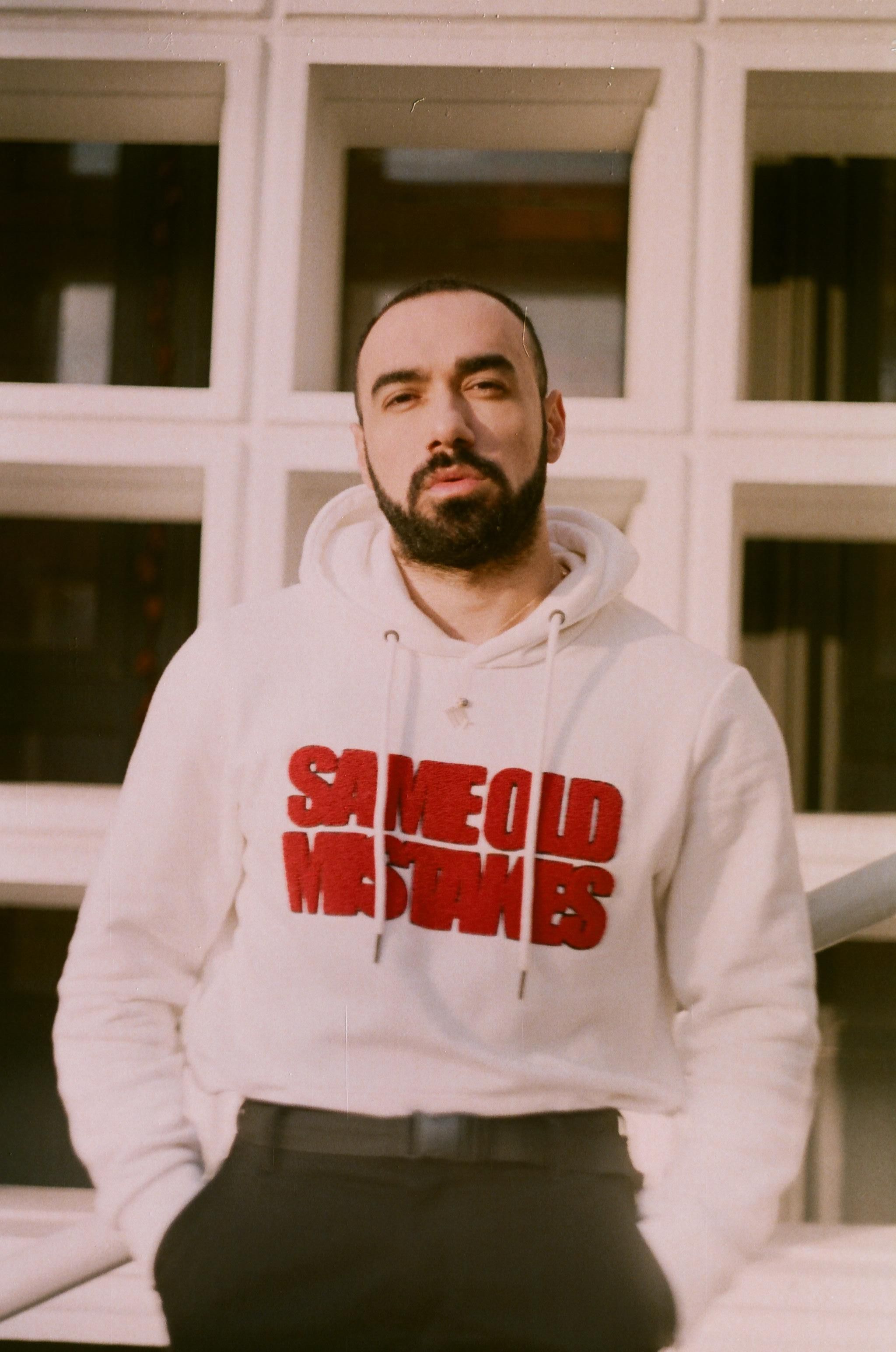 A man with a beard wearing a trendy white hoodie with text, posing outdoors against a modern building facade.