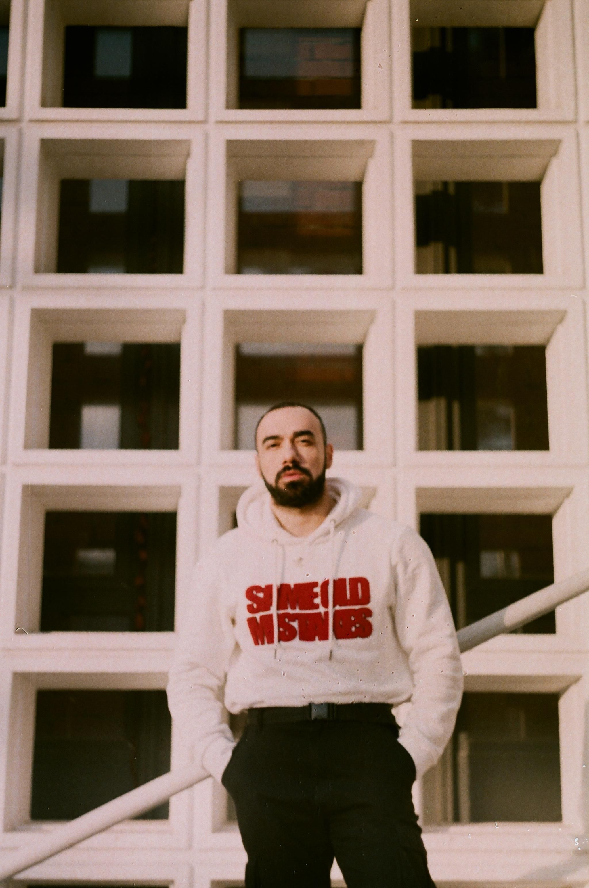 Young bearded man poses in front of modern architecture wearing a sweatshirt.