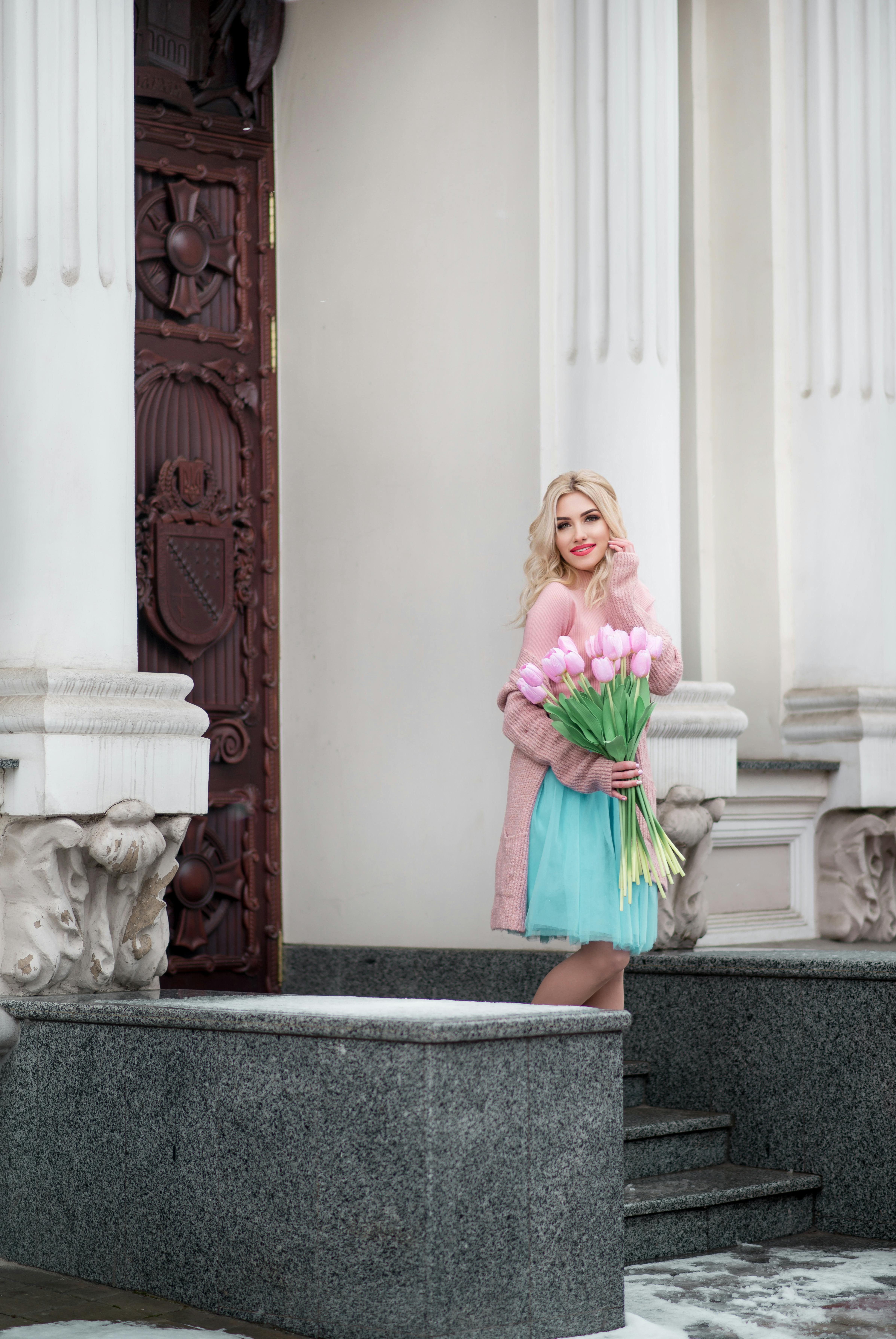 A stylish blonde woman in a dress holds a bouquet of tulips, posing by a decorative building.