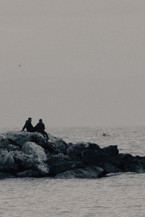 Free Two people sit on rocky shore overlooking the calm sea on a cloudy day. Stock Photo