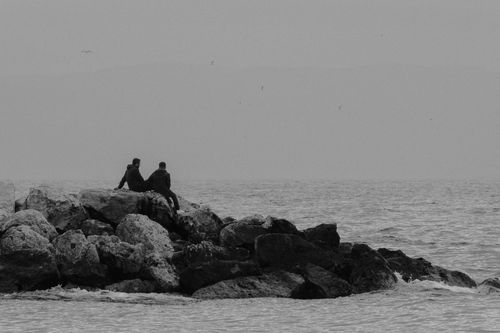 Free Two people sitting on seaside rocks, enjoying a peaceful black and white seascape. Stock Photo