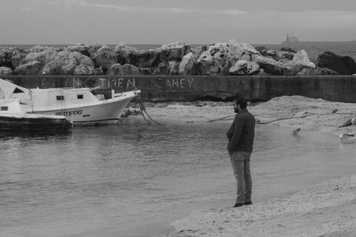 Free A man standing on a beach next to a moored boat, gazing at the sea, in black and white. Stock Photo
