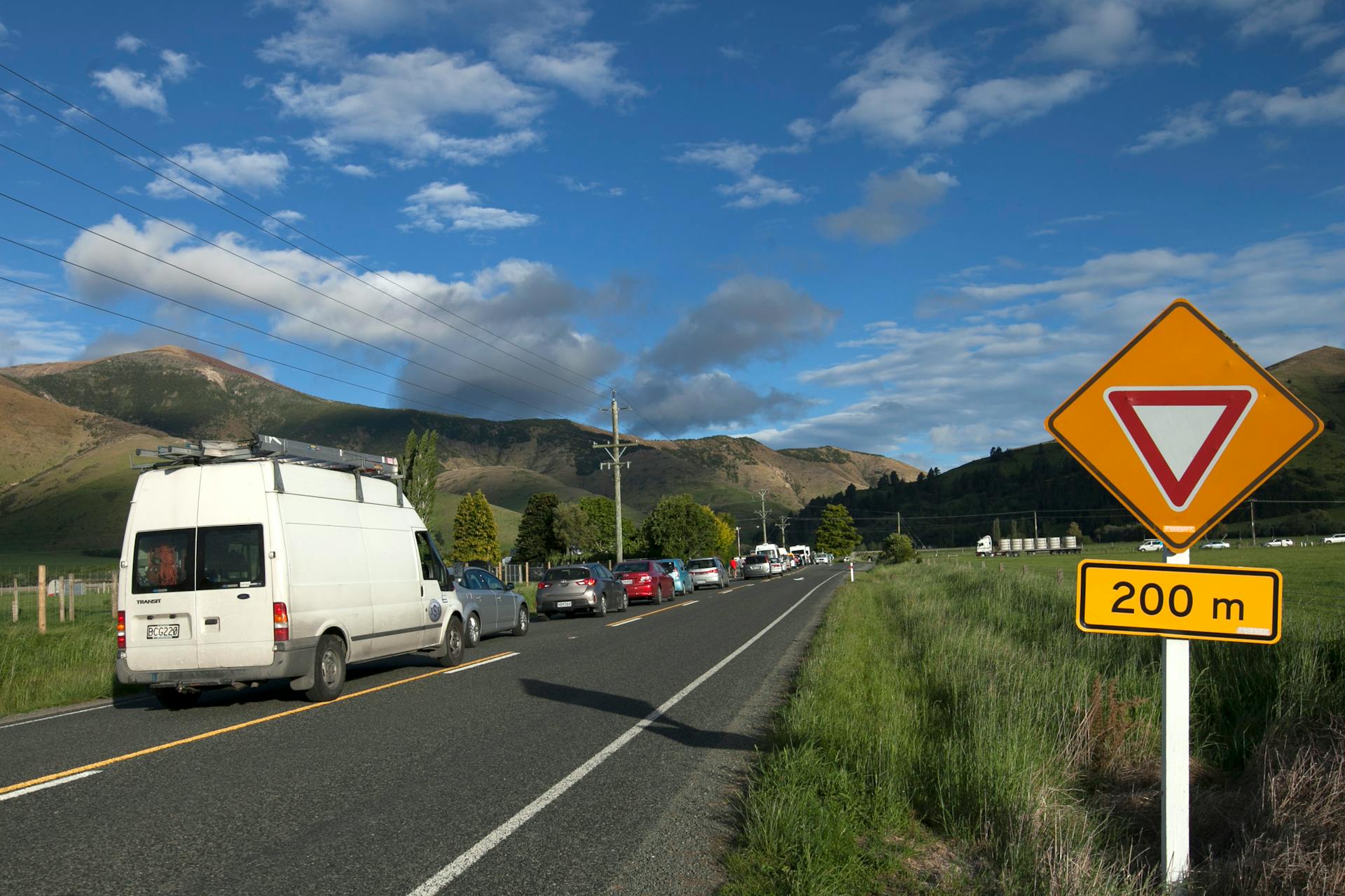 Traffic lined up on a rural road with a yield sign against a mountain backdrop.