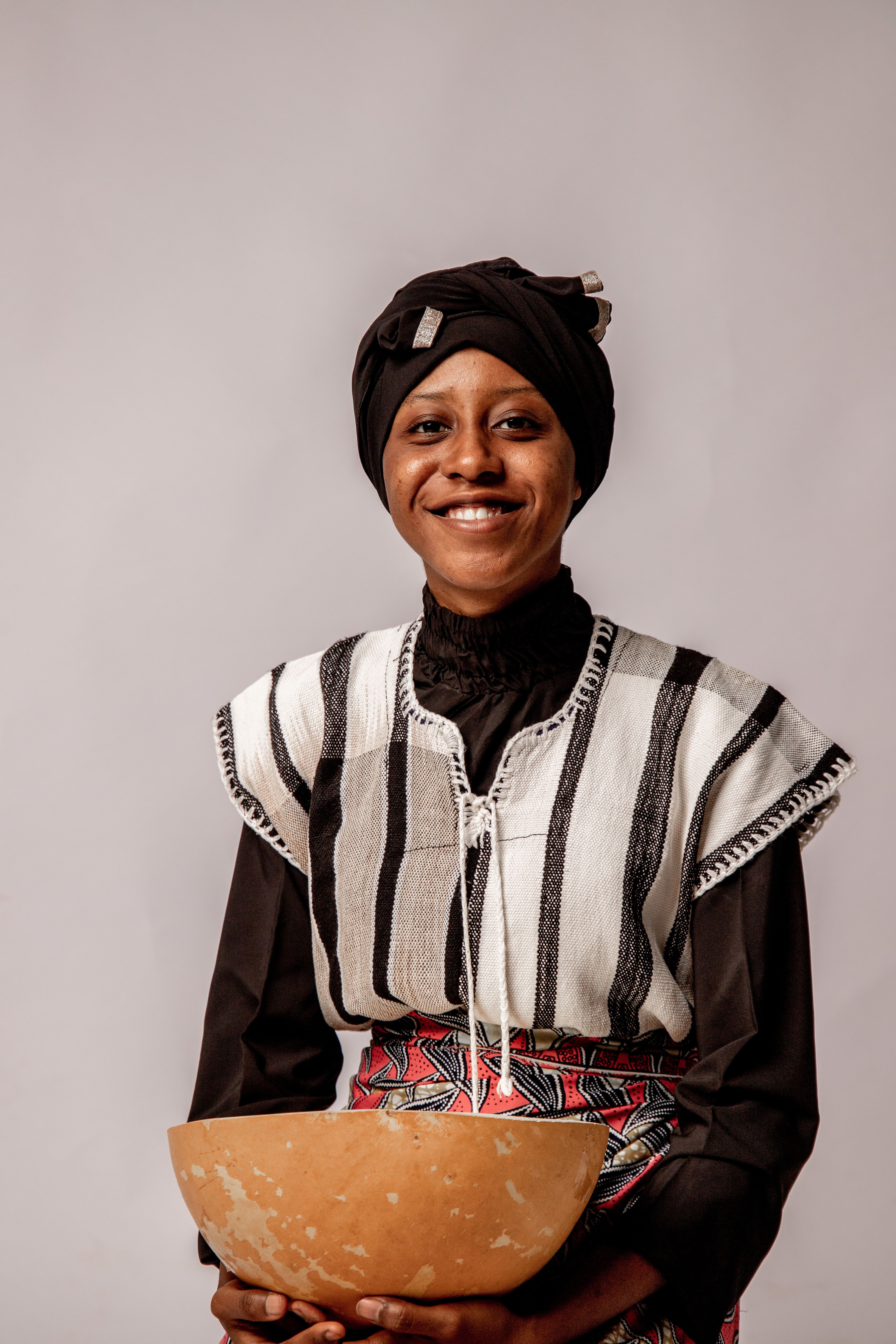 Smiling woman in traditional attire holding a pot, featuring vibrant patterns.