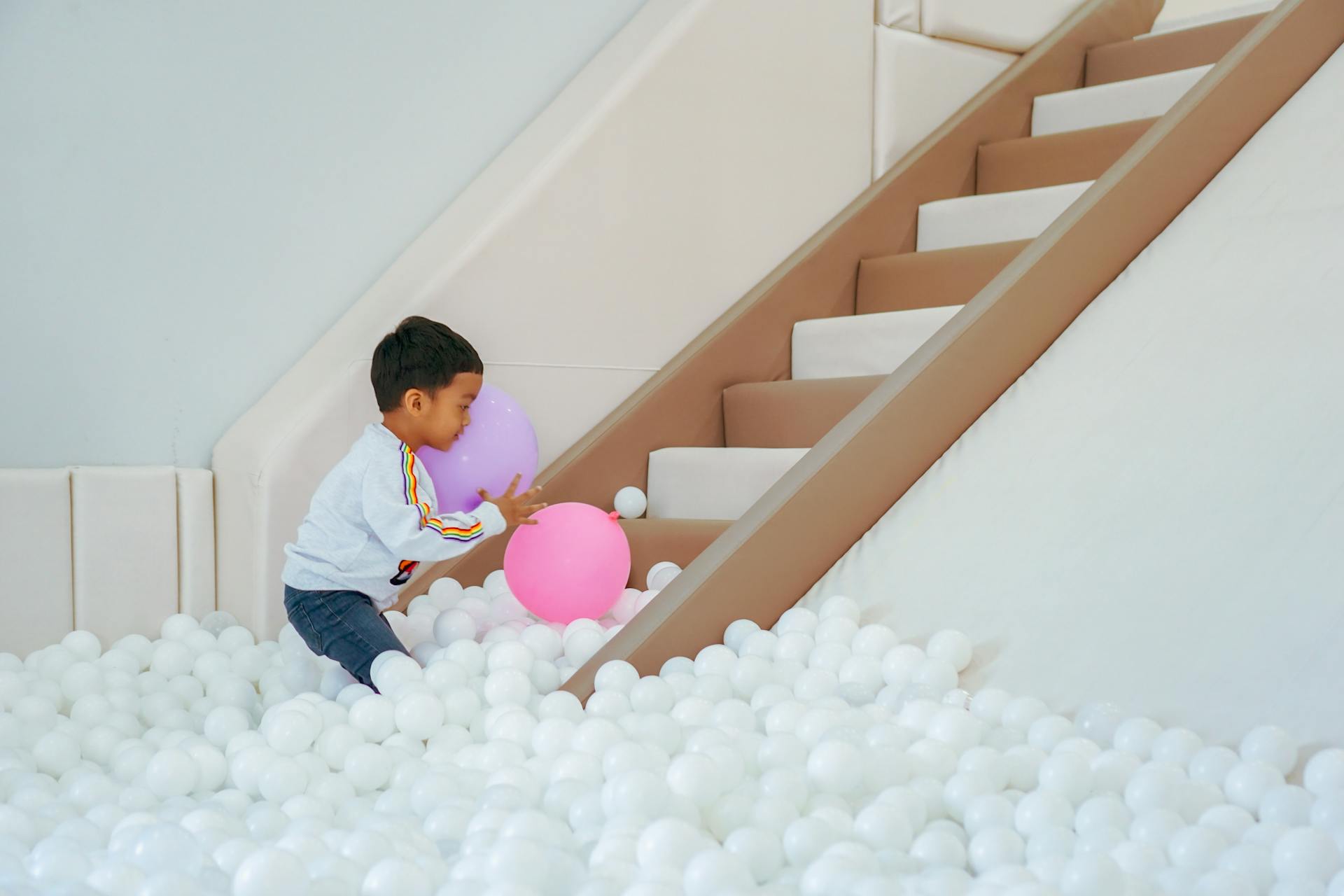 A young boy playing with colorful balloons on an indoor slide surrounded by a sea of white balls.