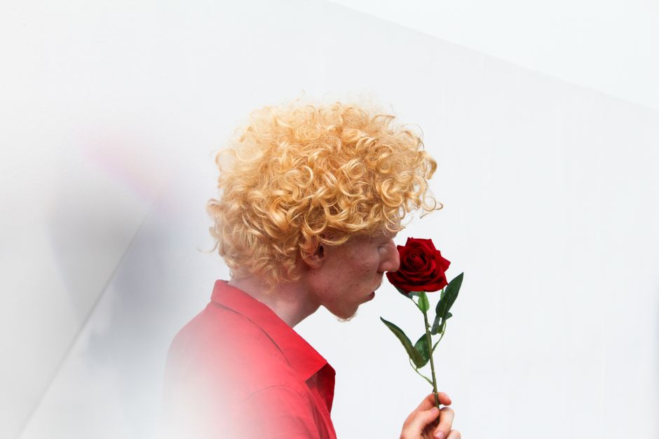 A man with curly blond hair holds a red rose, set against a minimalist white background.