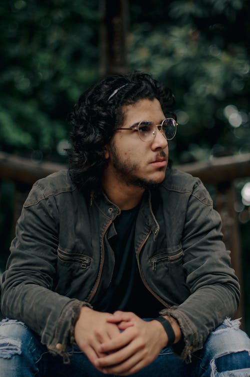 Young Man with Curly Hair and Eyeglasses Sitting Outside and Looking Away 