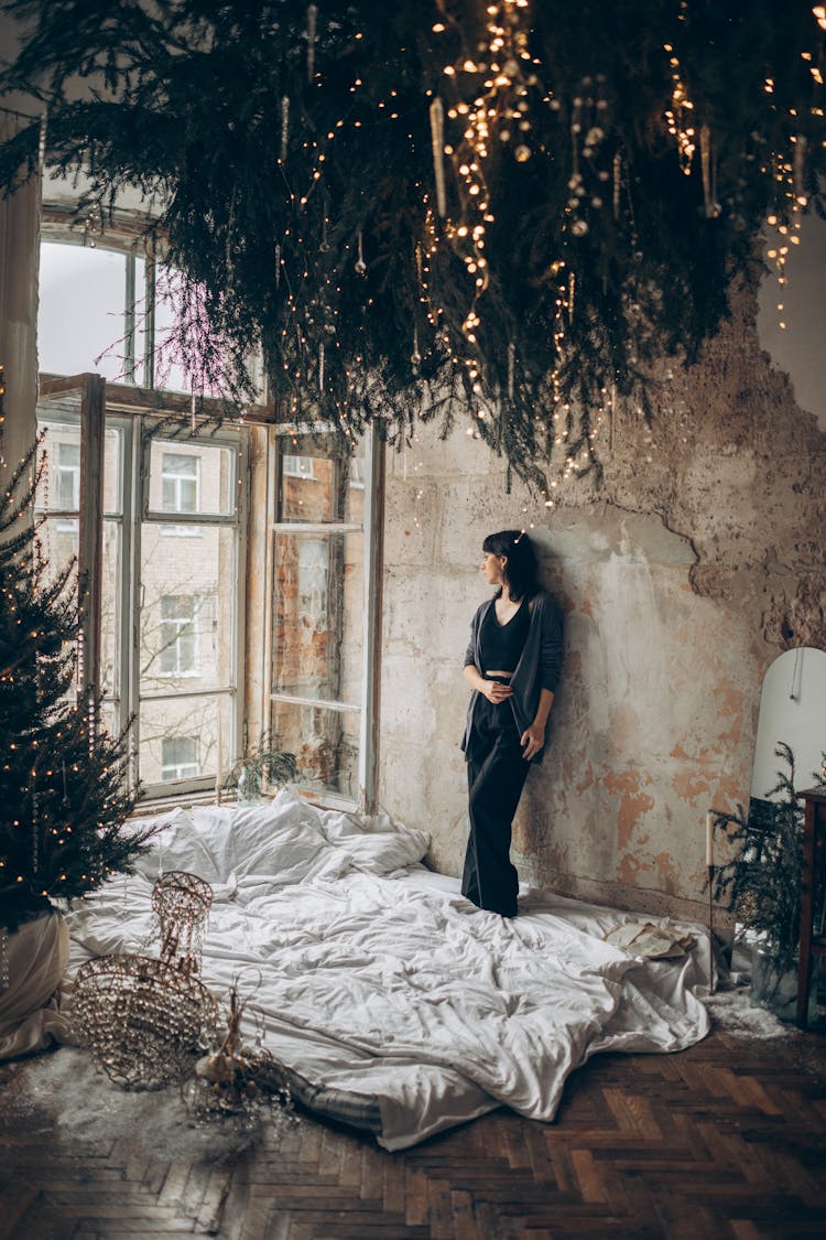 Woman Standing On A Bed And Looking Out The Window Of A Ruined Room Decorated For The Christmas