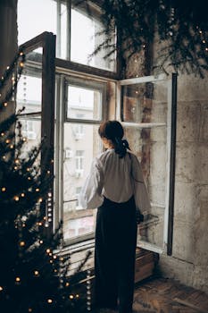A woman stands by a window in a cozy room with winter decorations.