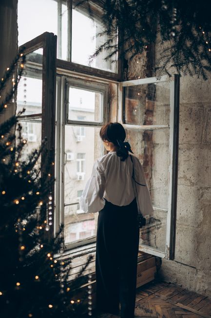 A woman stands by a window in a cozy room with winter decorations.