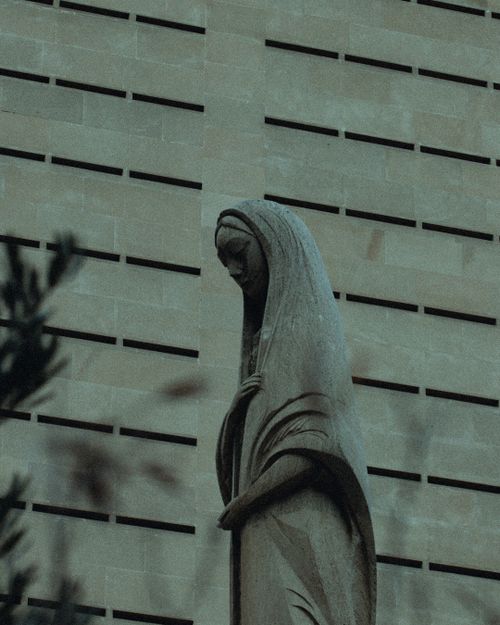 Free A serene statue of the Virgin Mary stands against a modern urban facade, depicting religious art in a city context. Stock Photo
