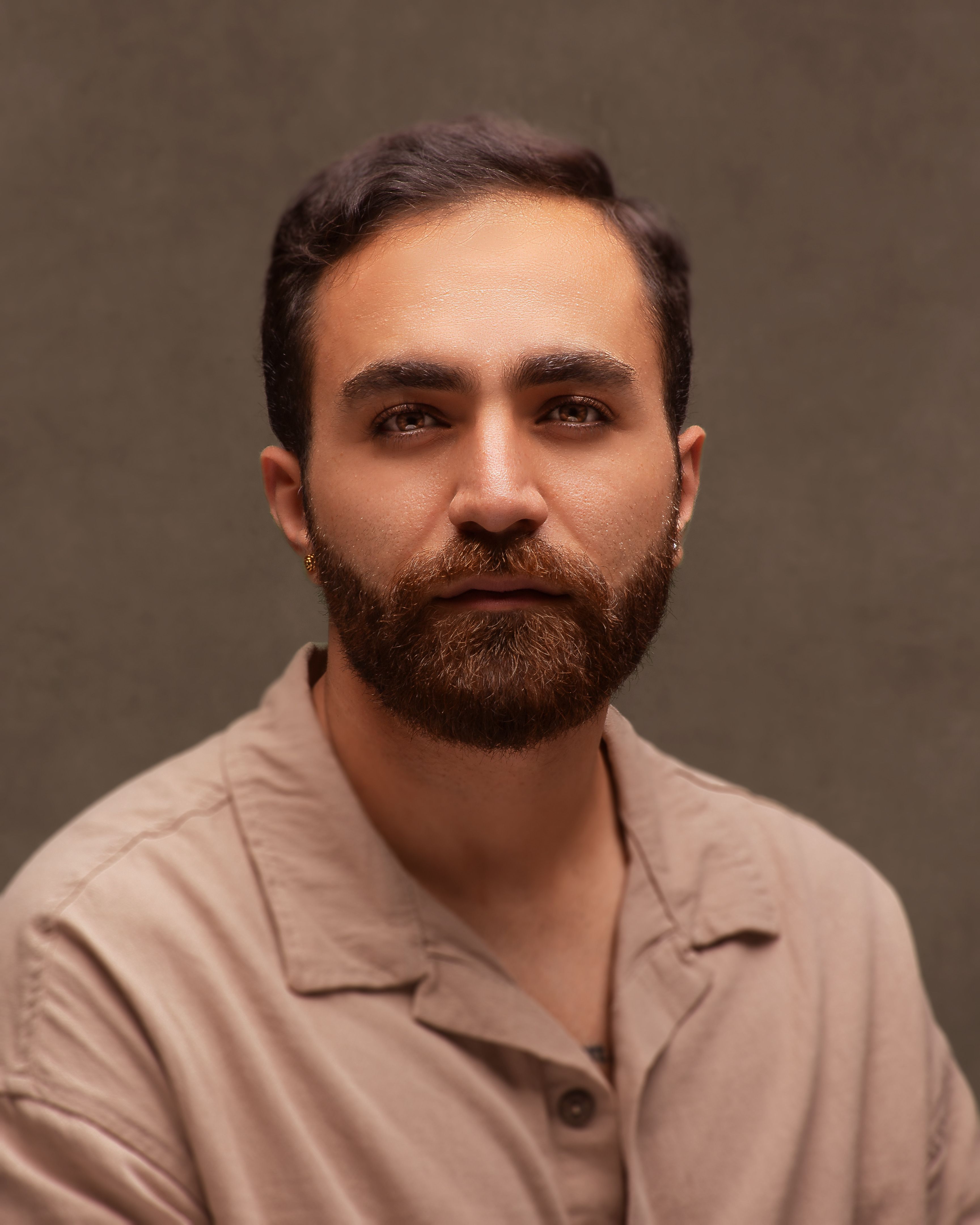 Studio portrait of a bearded adult man wearing a beige shirt, looking directly at the camera.