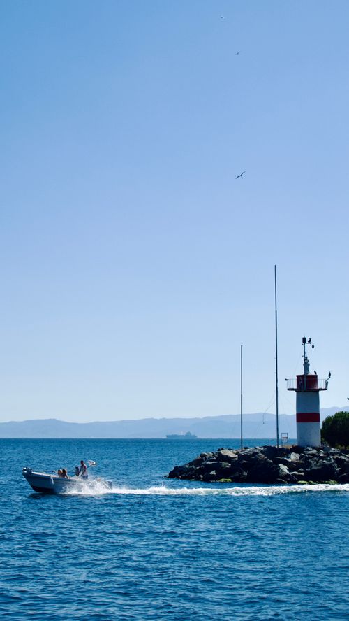 Free A motorboat speeds past a lighthouse by the sea under a clear blue sky. Stock Photo
