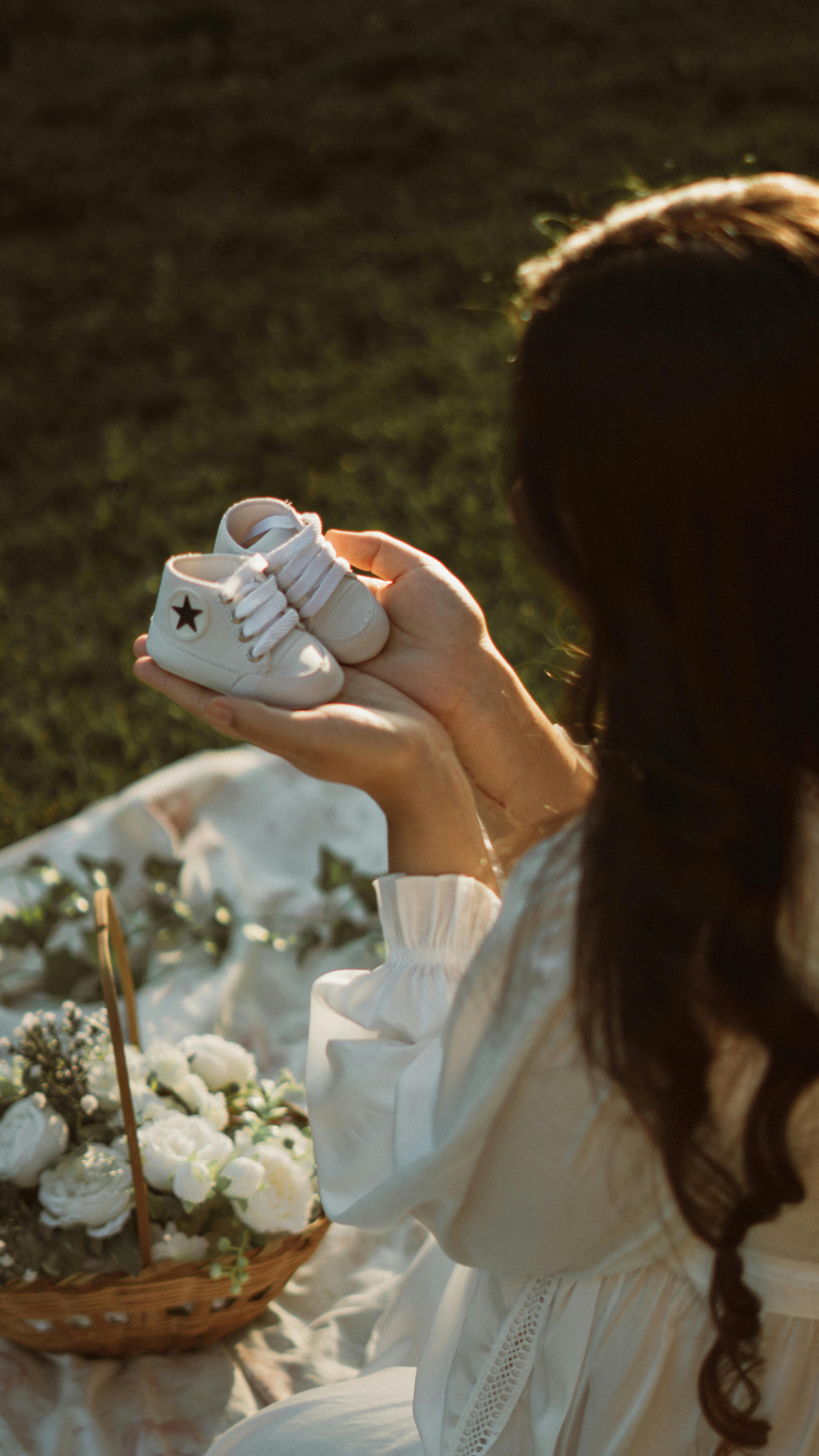 A woman lovingly holds baby shoes in a tranquil garden setting with white roses.