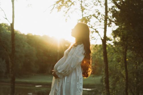 Free Pregnant woman in white dress enjoying a sunset in a serene forest setting. Stock Photo