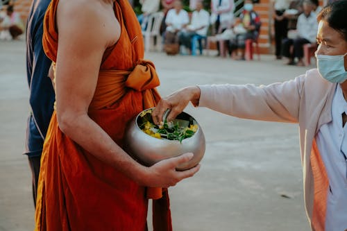Woman in Face Mask Reaching to Bowl Held by a Buddhist Monk