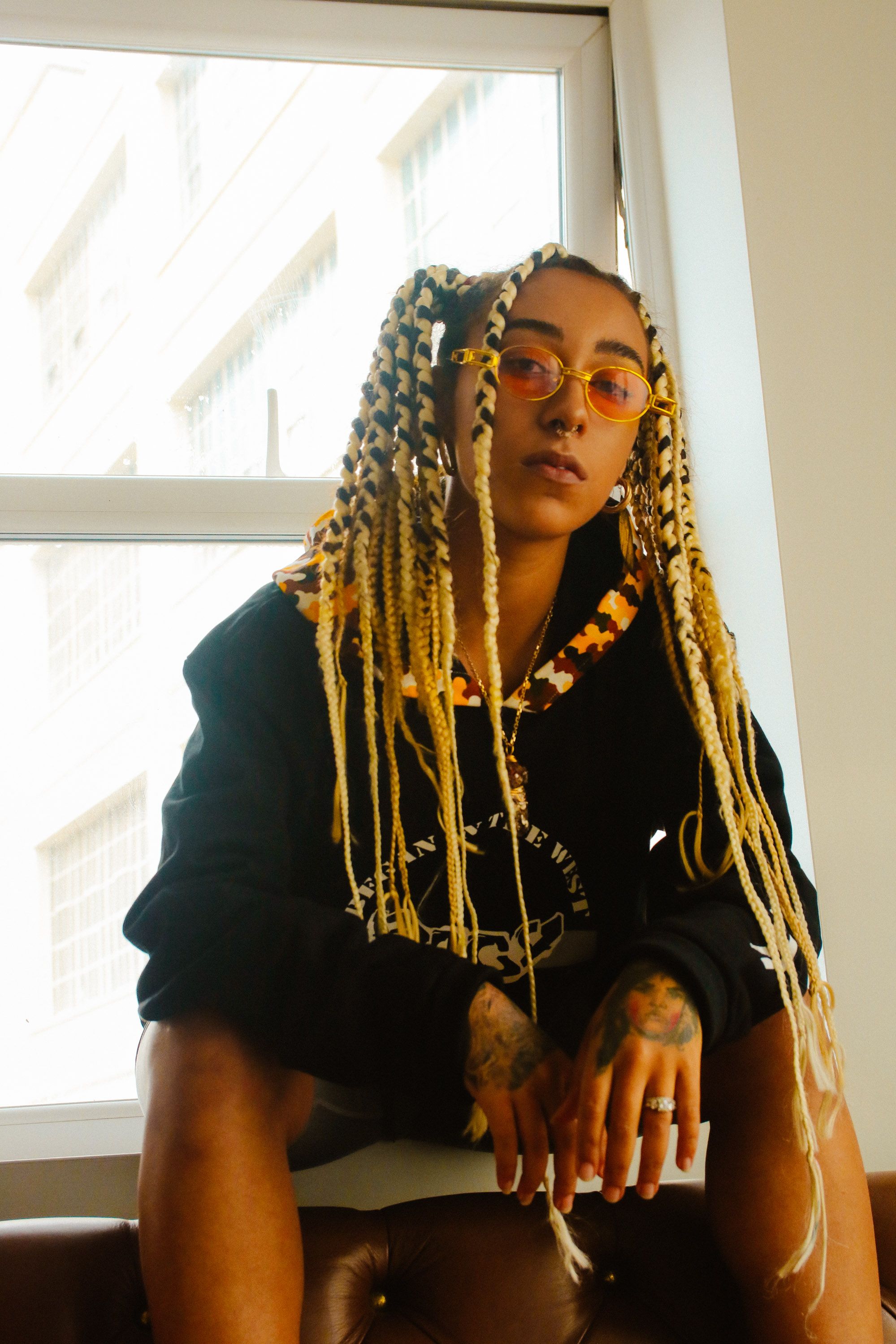 Stylish woman sitting by window in hoodie with colorful braids and glasses.