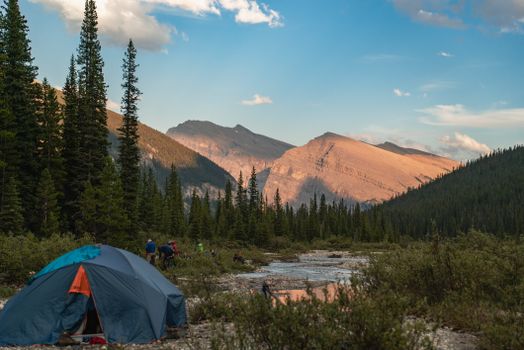 A serene campsite with a blue tent beside a river, surrounded by mountains and coniferous trees.
