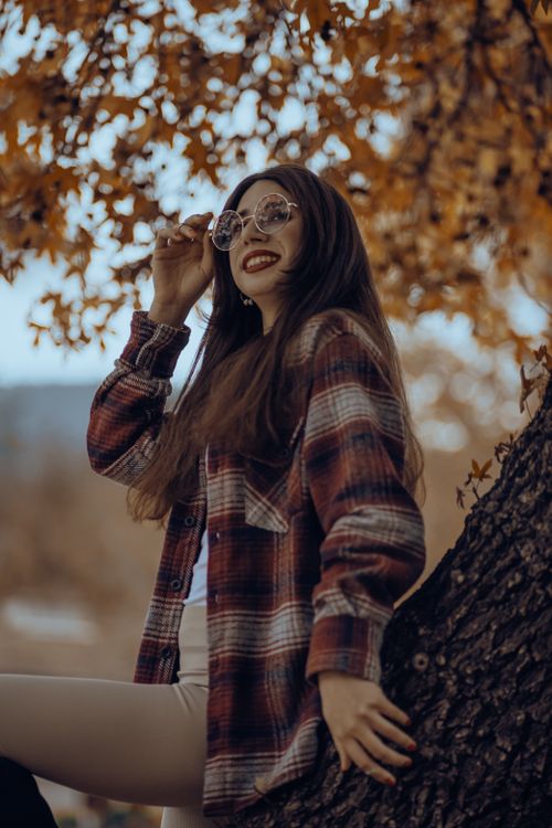 Free Smiling woman in plaid jacket and sunglasses leaning on tree outdoors. Stock Photo