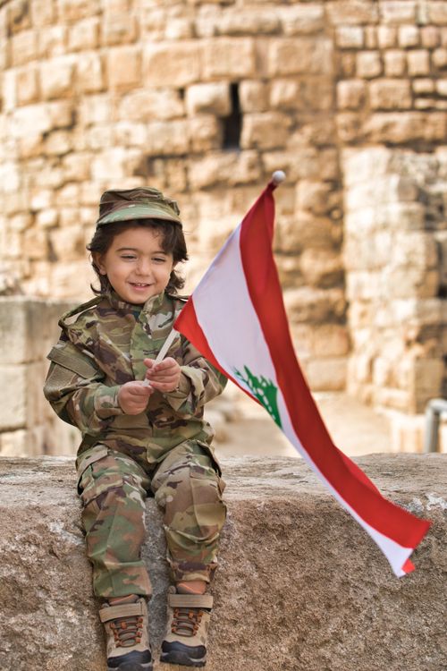 Free Smiling child in camouflage uniform holding Lebanese flag, expressing patriotism. Stock Photo