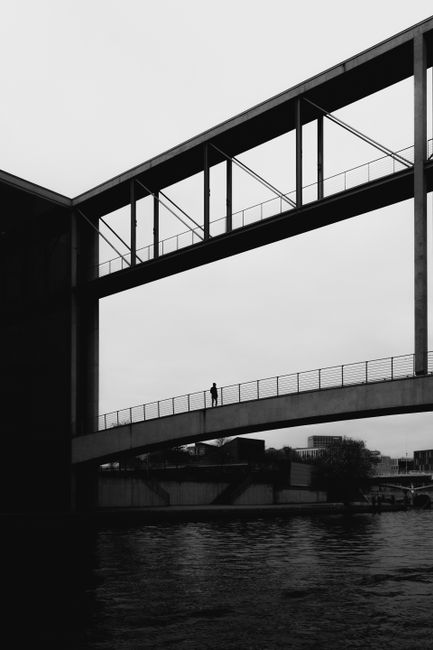 A lone figure walking across a modern footbridge over a river in an urban setting, captured in black and white.