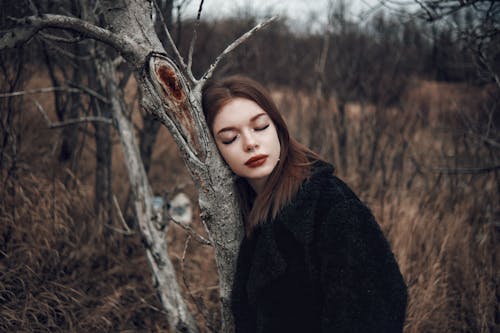 Beautiful Young Woman in a Coat Leaning Against a Leafless Tree