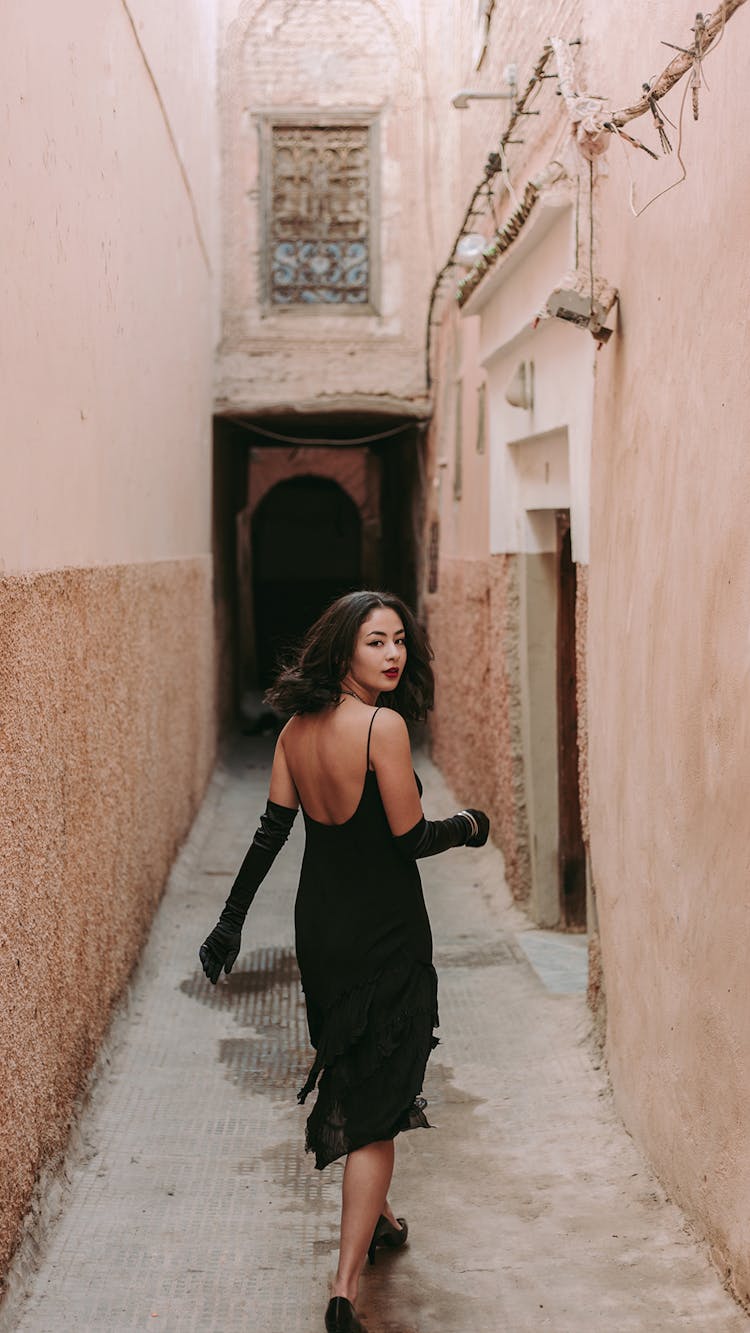 Elegant Woman In A Black Dress Walking In A Narrow Alley And Looking Over The Shoulder 