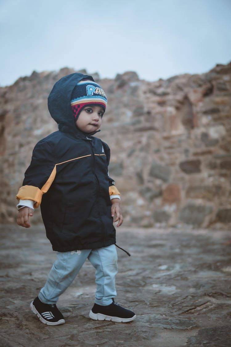 A Toddler In A Jacket And Hat Walking Outside 